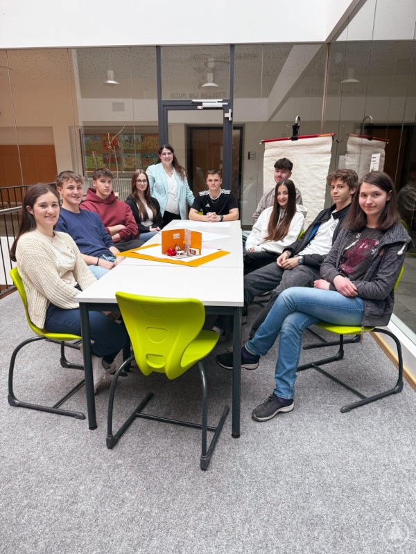 Die Schülerinnen und Schüler stellten im Gespräch mit Kulturreferentin Gabriela Rauscher ihre bisher erarbeiteten Inhalte und die bereits gestalteten Ausstellungselemente vor und erhielten Tipps von der Museumsleiterin zum Aufbau der Ausstellung und zur Gestaltung der Führungen. Eine Gruppe von Schülerinnen und Schülern sowie eine Frau stehen bzw. sitzen in einem modernen Raum um einen Tisch, im Hintergrund Flipcharts und Glaswände.