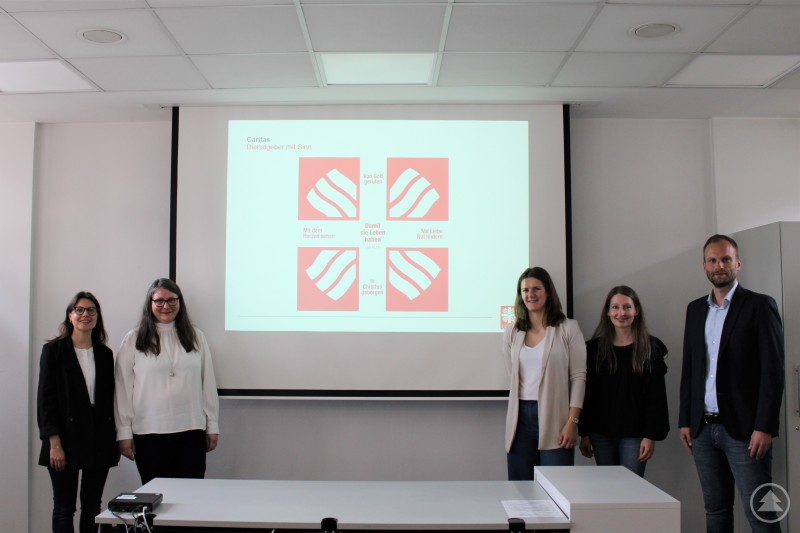 Sandra Ciupinski, Ursula Sendlinger, Tanja Ohrhallinger, Andrea Bayer und Thomas Furthmeier (v. l.) beim Austausch über neue Perspektiven der Zusammenarbeit zwischen der Caritas Passau und der Agentur für Arbeit. Fünf Personen stehen vor einer Leinwand in einem Konferenzraum. Auf der Leinwand ist das Caritas-Logo in vier Varianten mit Slogans wie „Mit dem Herzen sehen“ und „Von Gott gerufen“ zu sehen. Die Personen lächeln freundlich in die Kamera.