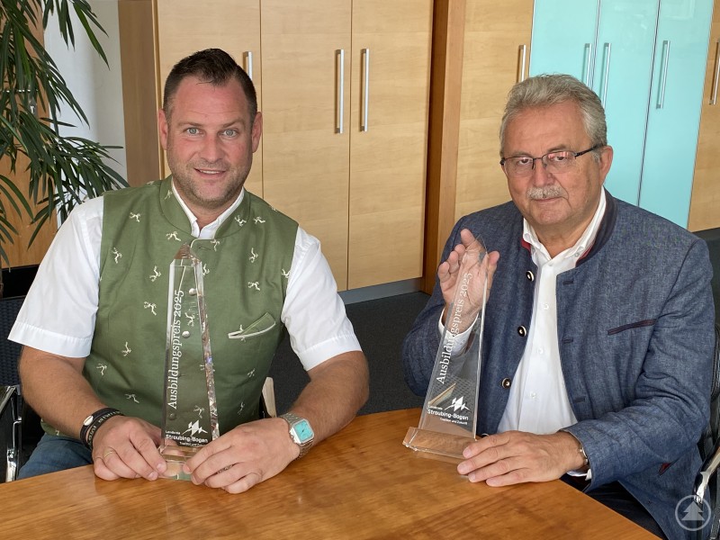 Wirtschaftsförderer Martin Köck (links) und Landrat Josef Laumer mit dem Ausbildungspreis des Landkreises Straubing-Bogen – Bewerbungen für 2025 sind ab sofort möglich. Zwei Männer in Tracht sitzen an einem Tisch und halten transparente Trophäen in der Hand.