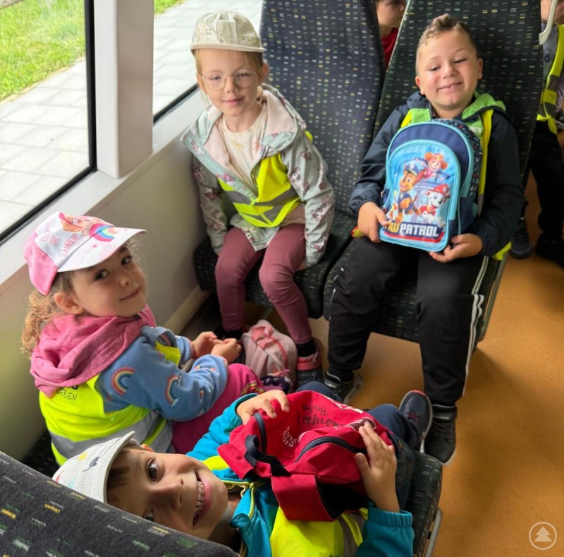 Mit Bus und Bahn ging es in die Wildnis hinein und über einen Pommes-Stopp wieder nach Hause. Eine Gruppe fröhlicher Kinder in Warnwesten sitzt in einem Zugabteil, einige mit bunten Rucksäcken.