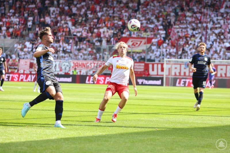 Das Bild zeigt eine Spielsituation zwischen dem SSV Jahn Regensburg (in Weiß-Rot) und dem MSV Duisburg (in Schwarz-Blau), bei der ein Regensburger Spieler den Ball in der Luft fixiert, während Gegenspieler in der Nähe sind. Im Hintergrund sind zahlreiche Fans und rot-weiße Banner zu sehen.