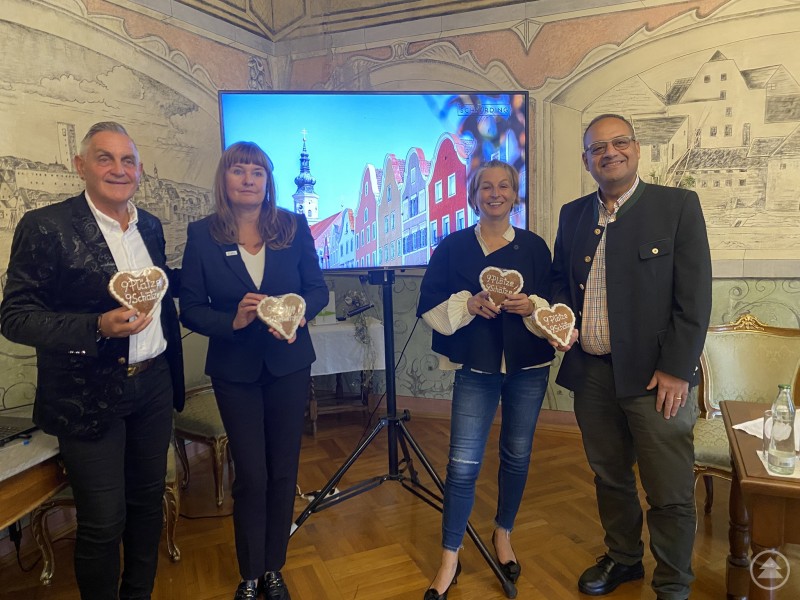 Bürgermeister Günter Streicher, Bettina Berndorfer, Wirtschaftsstadträtin Sandra Hofbauer und Maher Azer (v.l.n.r.) Vier Personen stehen in einem stilvoll dekorierten Raum mit Wandmalereien. Sie halten Lebkuchenherzen mit der Aufschrift „9 Plätze 9 Schätze“ in den Händen. Im Hintergrund ist ein Bildschirm mit einem Bild der Schärdinger Altstadt zu sehen.