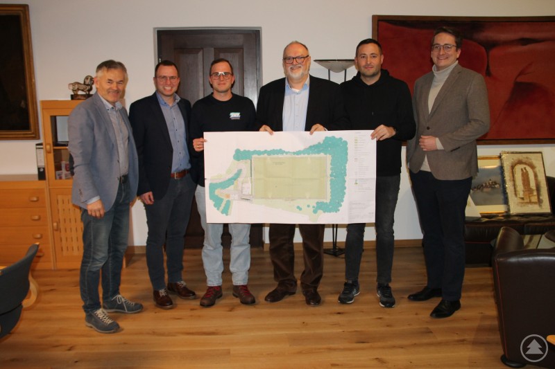 Oberbürgermeister Jürgen Dupper (3. von rechts) stellte zusammen mit Wirtschafts- und Bildungsreferent Werner Lang (links) und im Beisein von Stadtrat und Ehrenamtlichen Verwaltungsrat der Sportanlage Jonas Weidenthaler (rechts) die Planungen des Kunstrasenplatzes den Vereinsfunktionären des SC Batavia 72 Passau e. V. mit Sebastian Scharinger (2. von links), Fabian Scharinger (3. von links) und Aron Csizmadia (2. von rechts) vor. Sechs Männer stehen in einem Raum und halten gemeinsam einen großen Bauplan für einen neuen Sportplatz. Im Hintergrund sind Büromöbel und Bilder zu sehen.