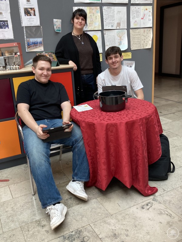 Quiz, Ausstellung und Mitmachaktionen sorgten in der Aula für ein buntes Treiben. Drei Jugendliche sitzen und stehen an einem Tisch mit roter Decke und Quizunterlagen. Vor ihnen steht ein schwarzer Lostopf.