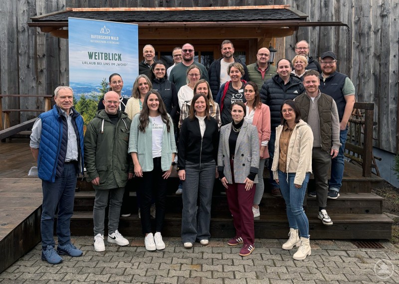 Die Tourismusschaffenden des Landkreises Freyung-Grafenau vor dem Brennereistadel der WoidHoamat in Schönbrunn am Lusen. Gruppe von rund 20 Tourismusakteuren steht vor einem rustikalen Holzgebäude und posiert gemeinsam für ein Gruppenfoto im Außenbereich.