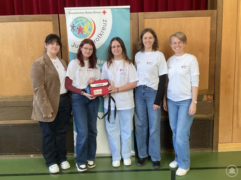 Die Schulsanitäterinnen überzeugten beim JRK-Regionalentscheid in Schönberg mit Teamgeist und starkem Einsatz. Fünf junge Frauen stehen vor einem Banner des Bayerischen Roten Kreuzes Jugendrotkreuz und präsentieren gemeinsam ihre Ausrüstung nach dem Regionalentscheid.