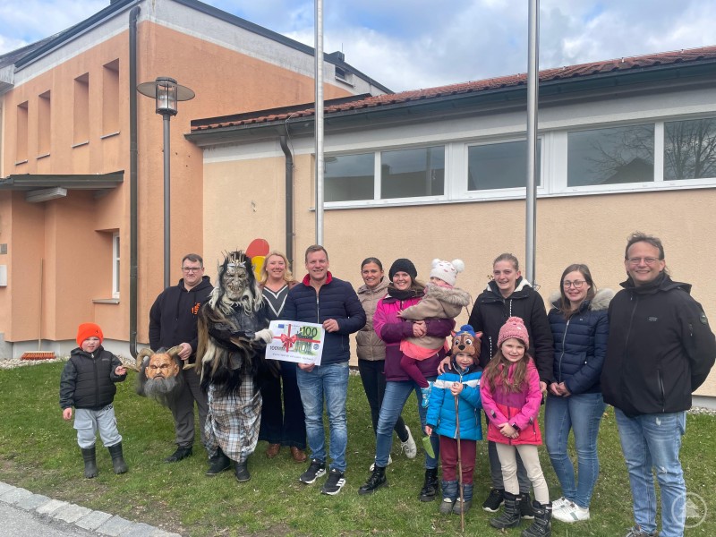 Die Lusengschwerl übergeben eine Spende an den Förderverein des Kindergartens Hl. Familie. Eine Gruppe von Erwachsenen und Kindern steht vor einem Gebäude, darunter eine Person in traditioneller Maskierung. In der Mitte wird ein Spendenscheck überreicht.