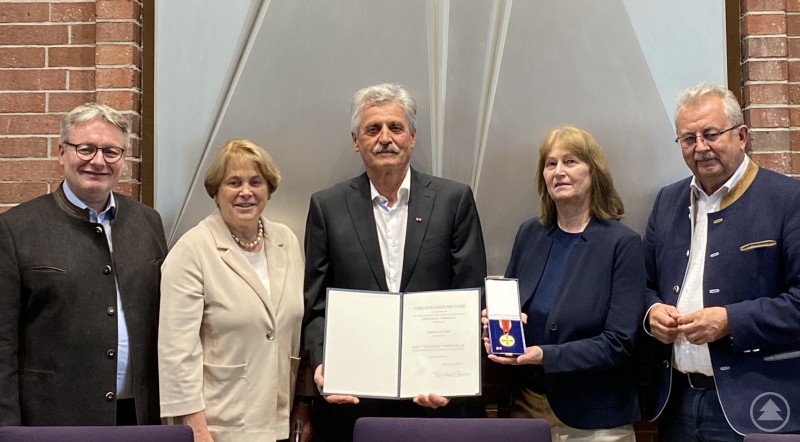 Landrat Josef Laumer überreichte Johann Berr im Beisein seiner Frau Christa, Bürgermeisterin Barbara Unger und MdL Josef Zellmeier die Verdienstmedaille der Bundesrepublik Deutschland. Fünf Personen stehen nebeneinander vor einer Wand mit dem bayerischen Staatswappen. Die mittlere Person hält eine Urkunde, eine danebenstehende Frau zeigt die Bundesverdienstmedaille in einer Box.