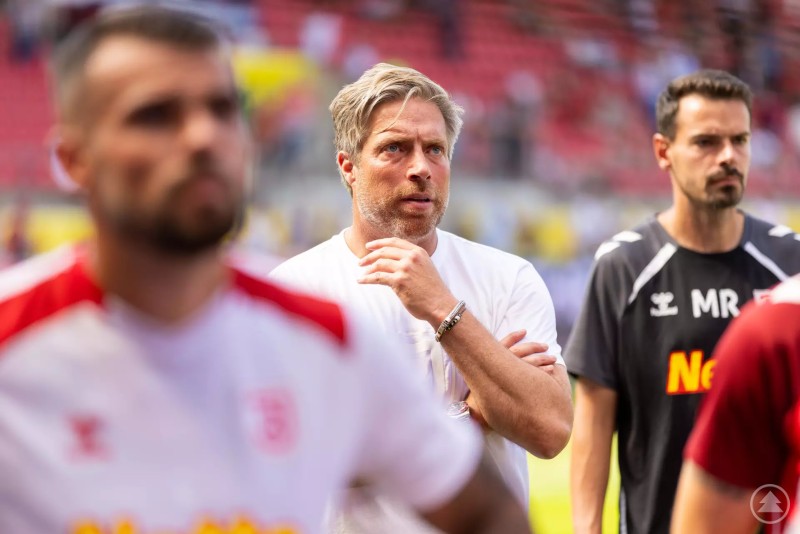 Das Bild zeigt eine Szene am Spielfeldrand eines Fußballstadions. Im Fokus steht ein Mann mit graublondem Haar und Bart, der ein weißes T-Shirt trägt und nachdenklich wirkt. Im Vordergrund und Hintergrund sind unscharf weitere Personen in Sportkleidung zu sehen, vermutlich Spieler oder Betreuer.