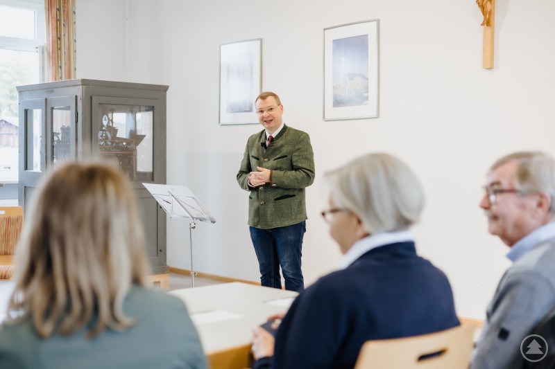 Ein Mann in traditioneller Tracht spricht vor einer kleinen Zuhörergruppe in einem hellen Raum.