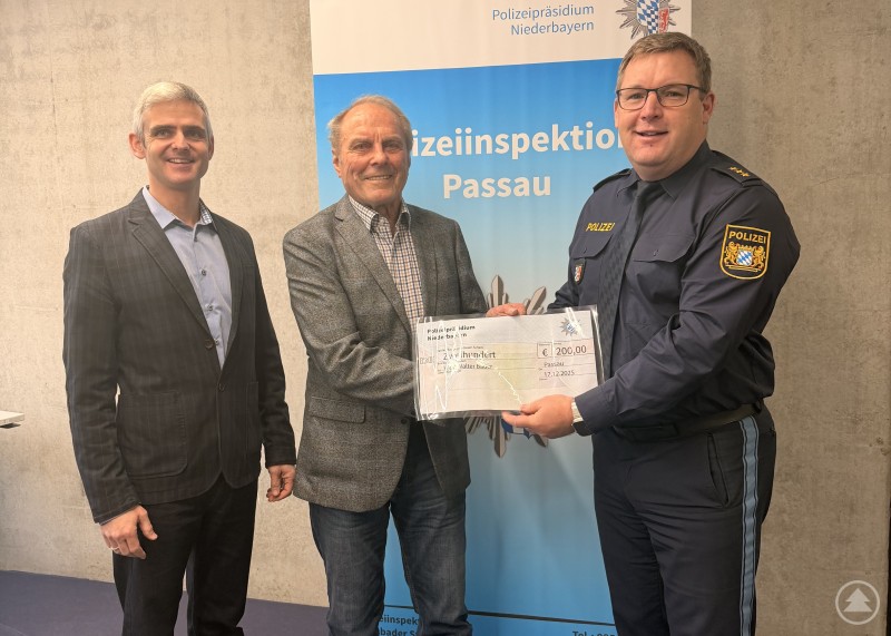 Kriminalrat Johannes Schimpfhauser, Walter Bauer und Polizeidirektor Christian Dichtl bei der Würdigung des couragierten Bürgers in Passau. Drei Männer stehen nebeneinander in einem Innenraum vor einem Roll-up der Polizeiinspektion Passau. In der Mitte hält ein älterer Mann ein Schreiben in den Händen, flankiert von zwei Polizeiführungskräften in Zivil und Uniform.
