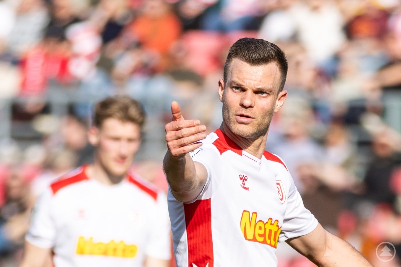 Benedikt Saller im Spiel gegen den SSV Ulm. Ein Spieler des SSV Jahn Regensburg im weiß-roten Trikot zeigt während eines Spiels mit ausgestrecktem Arm eine dirigierende Geste auf dem Spielfeld, im Hintergrund sind Zuschauer und ein weiterer Spieler unscharf zu erkennen.