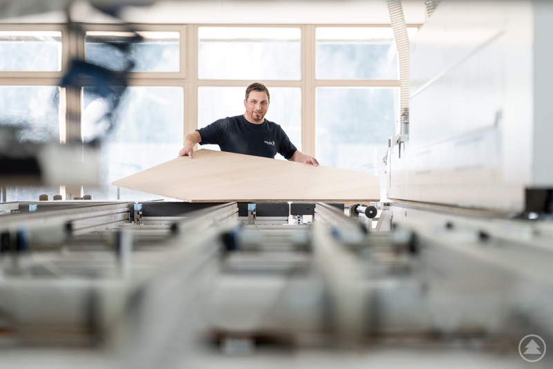 Ein Mitarbeiter legt in einer modernen Holzverarbeitungsmaschine eine gro&szlig;e Holzplatte ein. Die Szene zeigt eine pr&auml;zise CNC-Bearbeitung in einer hellen Produktionshalle.