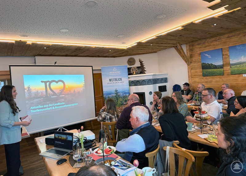 Vortragssituation in einem gem&uuml;tlichen Gastraum, mehrere Personen sitzen an Tischen und verfolgen eine Pr&auml;sentation auf einer Leinwand.