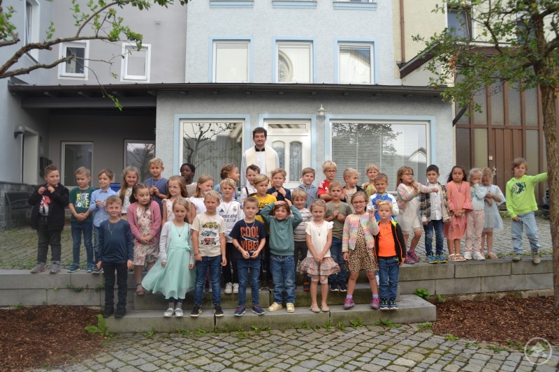 Vor der Kirche gab es erst Seifenblasen gen Himmel und dann ein Gruppenbild der ABC-Anwärter mit Pfarrer Simon Steinbauer. Eine große Gruppe von Kindergartenkindern posiert mit einem Pfarrer für ein Abschlussfoto vor einem Gebäude.