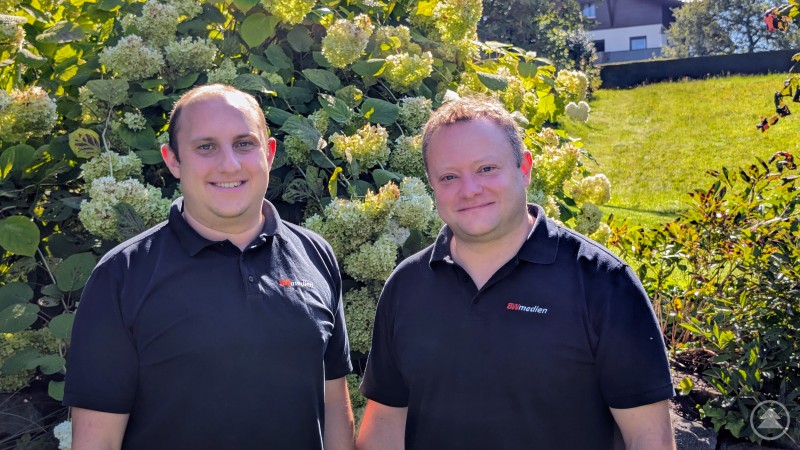 Die BWmedien-Geschäftsleitung im Doppelpack: Stefan Behringer (rechts) und Andreas Bildl (links) führen das Unternehmen mit Herz, Verstand und klarer Aufgabenteilung in die digitale Zukunft. Andreas Bild (links) und Stefan Behringer (rechts) stehen nebeneinander vor einem Strauch mit weißen Blüten. Beide tragen schwarze Poloshirts mit dem Logo BWmedien.