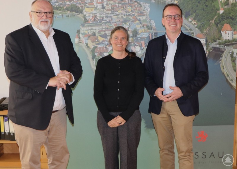 Oberbürgermeister Jürgen Dupper (li.) begrüßt den neuen Geschäftsführer des Niederbayern-Forums, Dr. Reinhard Saller, zusammen mit Heidemarie Bartl, Dienststellenleitung Wirtschaftsförderung, im Amtszimmer. Drei Personen stehen vor einem Wandbild der Stadt Passau, zwei Männer im Anzug flankieren eine Frau in schwarzer Kleidung.