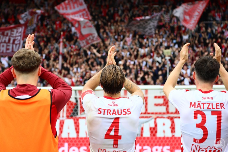 Die Jahn-Spieler bedanken sich nach dem 2:0-Heimsieg gegen den SC Verl bei den Fans für die lautstarke Unterstützung im Jahnstadion Regensburg. Fußballspieler des SSV Jahn Regensburg applaudieren gemeinsam mit den Fans. Im Hintergrund jubelt die große rot-weiße Fankurve mit Fahnen.