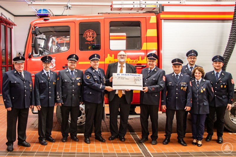 Oberbürgermeister Jürgen Dupper (5. v. links) nimmt den symbolischen Scheck von Vorstand Stefan Riegler (6. v. links) und dem 1. Kommandanten Hermann Obermüller (7. v. links) im Beisein der Vorstandschaft entgegen. Eine Gruppe von Feuerwehrleuten in Uniform steht in einer Reihe vor einem roten Feuerwehrfahrzeug. In der Mitte überreicht ein Mann im Anzug einen symbolischen Scheck, der von mehreren Personen gemeinsam gehalten wird.