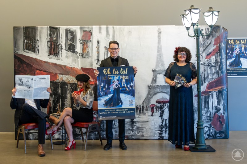 Foto v.l.n.r.: Johannes Winter, Bianca Pelzer, Oberbürgermeister Dr. Christian Moser und Kulturamtsleiterin Sabine Saxinger Vier Personen posieren vor einer künstlerischen Paris-Kulisse mit Eiffelturm. Zwei sitzen – einer mit Zeitung, eine im Pariser Look mit Baskenmütze und roter Tasse –, während Oberbürgermeister Dr. Christian Moser ein großes Veranstaltungsplakat hält. Rechts steht Kulturamtsleiterin Sabine Saxinger neben einer dekorativen Straßenlaterne.