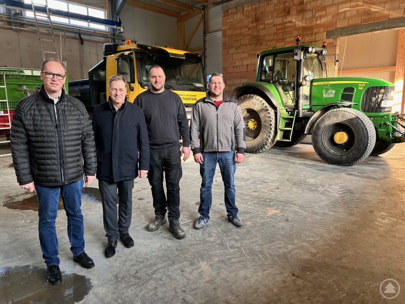Vier M&auml;nner stehen in einer gro&szlig;en Maschinenhalle vor einem Traktor und einem Lkw. Die Halle ist noch im Ausbau, die Fahrzeuge sind prominent platziert.