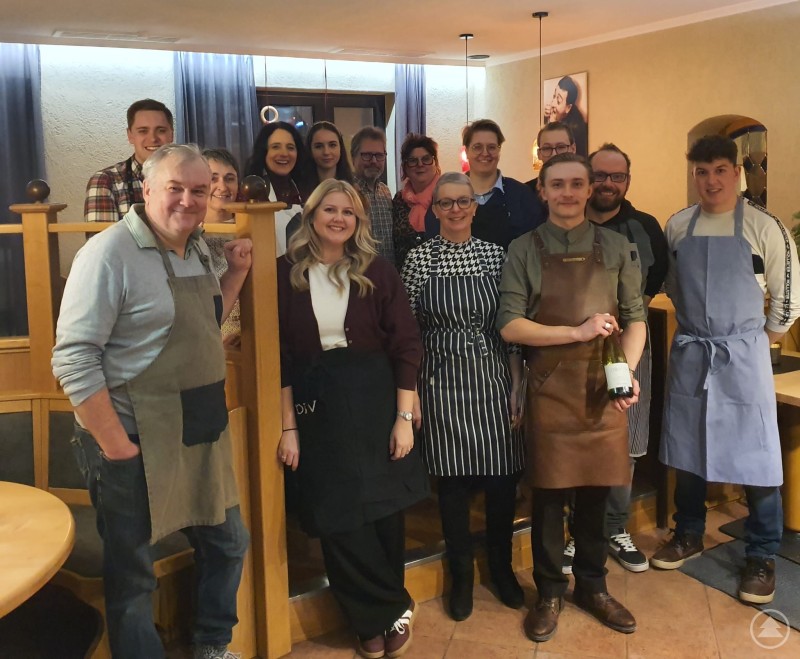 Das Kreativteam der Werbegemeinschaft Grafenau traf sich in der Pizzeria da Roberto zum Austausch und Pizzaworkshop. Gruppenfoto von rund 15 Personen in einem Restaurant, einige tragen Schürzen, im Vordergrund stehen zwei Personen mit Weinflasche und Schürzen, aufgenommen bei einem gemeinsamen Workshop.