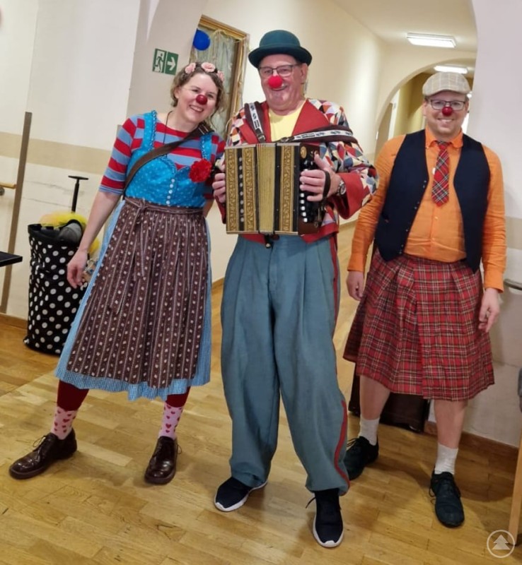 Die lustigen Clowns sorgten für jede Menge Unterhaltung Drei bunt gekleidete Clowns mit roten Nasen stehen im Flur eines Seniorenstifts, einer spielt Akkordeon.