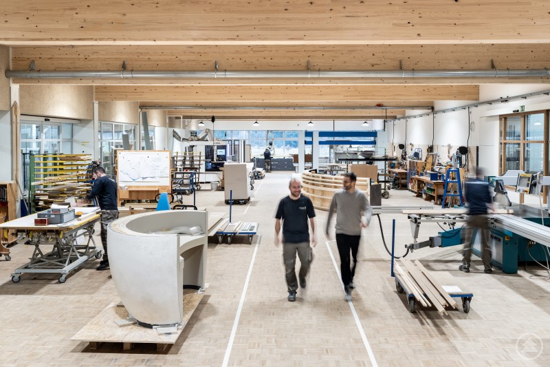 In der Werkstätte von GRAF entstehen hochwertige Innenausbau-Lösungen für Hotels, Gastronomie und Sonderprojekte. Blick in die großzügige Produktionshalle eines Holzverarbeitungsbetriebs. Mitarbeitende arbeiten an verschiedenen Maschinen und Werkbänken an Möbel- und Innenausbauprojekten.