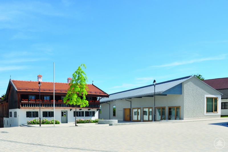 Maitenbeth, Lkr. Mühldorf am Inn (Oberbayern). Kirchplatz mit denkmalgeschützter Alter Post (links) und dem Ratssaal des Neuen Rathauses (rechts): Der Neubau des Rathauses nimmt die Maßstäblichkeit der Alten Post auf. Beide Gebäude schließen den Kirchplatz auf zwei Seiten räumlich ab. Über Fenstertüren ist der Ratssaal mit dem Freiraum des Kirchplatzes verbunden. Alt und Neu treten in einen gemeinsamen Dialog.