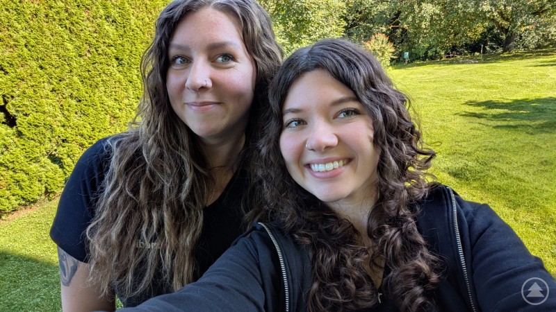 Kathleen Müller (Kathi) und Jana Spannbauer - die zwei Grinsekatzen haben immer gute Laune Zwei junge Frauen mit langen, lockigen Haaren posieren lachend für ein Selfie im Grünen. Im Hintergrund: dichter, grüner Rasen und eine hohe Hecke bei sonnigem Wetter.