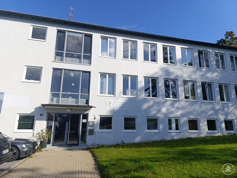 Das Bild zeigt ein modernes, helles Gebäude mit mehreren großen Fenstern und einem Haupteingang mit Glasfront. Links neben dem Eingang stehen geparkte Autos. An der Fassade ist ein Schild mit der Aufschrift „Berufsfachschule für Physiotherapie“ zu sehen. Das Gebäude steht in einer gepflegten Umgebung mit Rasenfläche im Vordergrund und blauem Himmel im Hintergrund.