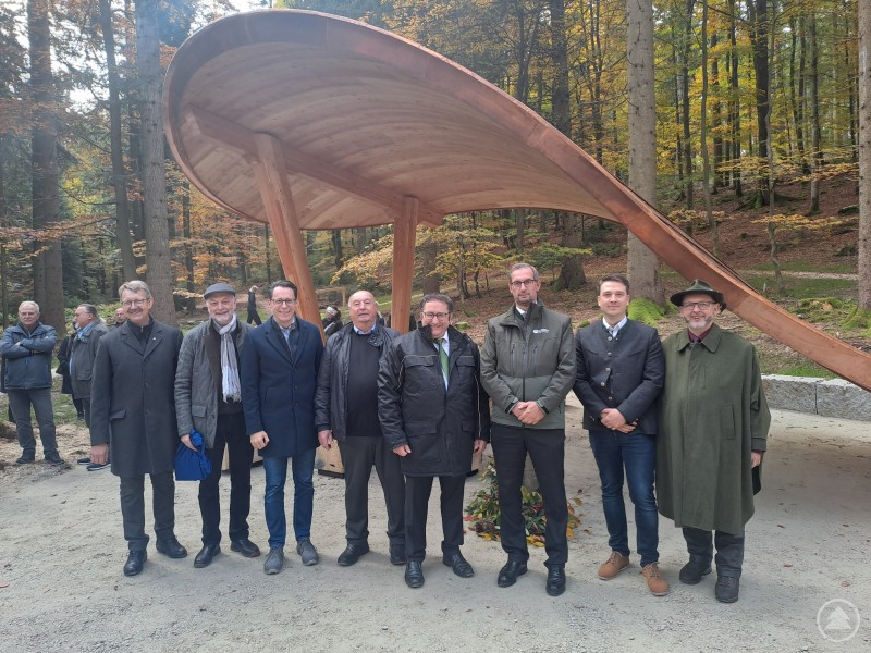 Freuen sich über die am 16.10. erfolgte Eröffnung des Stillen Waldes Rusel: Diakon Willi Poiger, Pfarrer Franz Deffner, Deggendorfs Oberbürgermeister Dr. Christian Moser, stellvertretender Landrat Josef Färber, Staatssekretär Tobias Gotthardt, BaySF-Vorstand Manfred Kröninger, Forstbetriebsleiter Markus Höhensteiger sowie dessen Amtsvorgänger Jürgen Völkl (v.li.). Acht Männer stehen nebeneinander vor einer modernen Holzüberdachung im Wald. Hinter ihnen sind herbstlich gefärbte Bäume zu sehen.