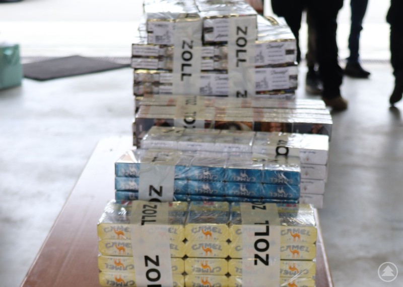 Vom Zoll sichergestellte Schmuggelzigaretten im Rahmen der Schwerpunktkontrolle der Grenzpolizei Passau. Mehrere gestapelte Stangen Zigaretten, teils mit Banderolen des Zolls versehen, liegen auf einem Tisch in einer Halle. Im Hintergrund sind unscharf Personen zu erkennen.