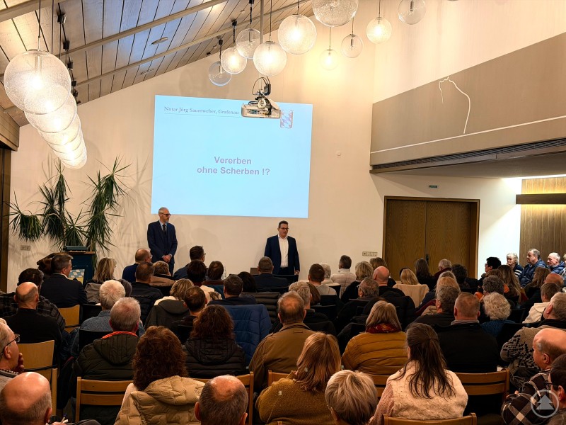 Zwei M&auml;nner im Anzug stehen vor einer gro&szlig;en Leinwand im B&uuml;rgersaal, w&auml;hrend zahlreiche G&auml;ste gespannt zuh&ouml;ren.