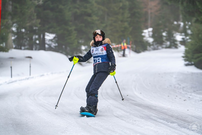 Ein junges Kind f&auml;hrt mit Skiern eine schneebedeckte Strecke bergab. Es tr&auml;gt eine Startnummer und wird von Zuschauern am Streckenrand beobachtet.