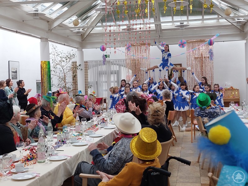 Viel Applaus erhielt die Büchlberger Faschingsgarde für ihren Auftritt Eine Kinder- und Jugendgarde führt einen akrobatischen Tanz in einem festlich geschmückten Saal auf, die Seniorinnen und Senioren applaudieren an gedeckten Tischen.
