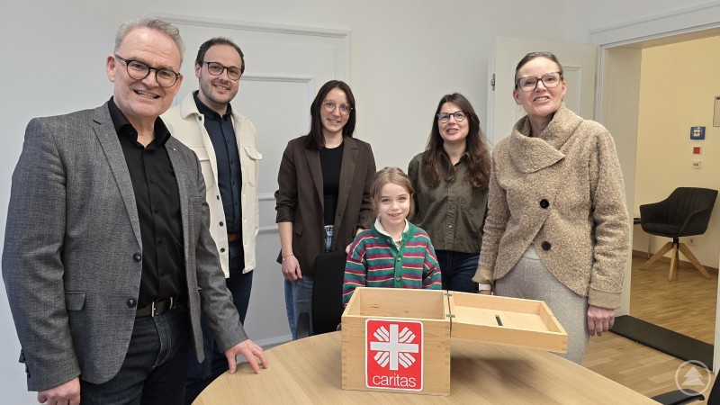 Da hatte die siebenjährige Charlotte ein glückliches Händchen. Die Caritas-Vorstände Andrea Anderlik und Stefan Seiderer sowie Patrik Lang, Melanie Jocham und Sandra Ciupinski von der Personalabteilung danken den Mitarbeiter:innen für ihre Spenden. Eine Gruppe von sechs Erwachsenen und einem Mädchen steht in einem Büro um einen Tisch mit einer Holzbox mit Caritas-Logo. Die Beteiligten lächeln in die Kamera, während das Mädchen als Losfee bei der Ziehung der Gewinner der Restcent-Aktion geholfen hat.