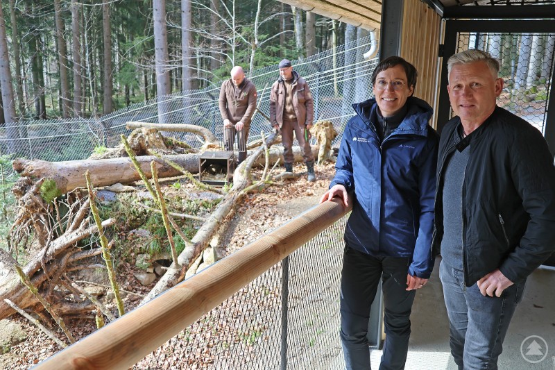 Nationalparkleiterin Ursula Schuster und Bürgermeister Alfons Schinabeck bei der Eröffnung des erweiterten Wildkatzen-Geheges im Tier-Freigelände. Zwei Personen stehen an einem Gehege im Tier-Freigelände und blicken auf eine naturnah gestaltete Anlage mit Holzstämmen und Waldkulisse, in der Wildkatzen leben.
