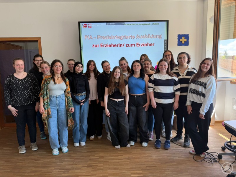 Die Auszubildenden des Sozialpädagogischen Einführungsjahres (SEJ) informierten sich aus erster Hand über die neue praxisintegrierte Ausbildung. Eine Gruppe junger Frauen steht lächelnd in einem Klassenzimmer vor einem Bildschirm, auf dem der Titel „PIA – Praxisintegrierte Ausbildung zur Erzieherin/zum Erzieher“ angezeigt wird. Im Hintergrund ist ein Fenster mit Blick nach draußen zu sehen.