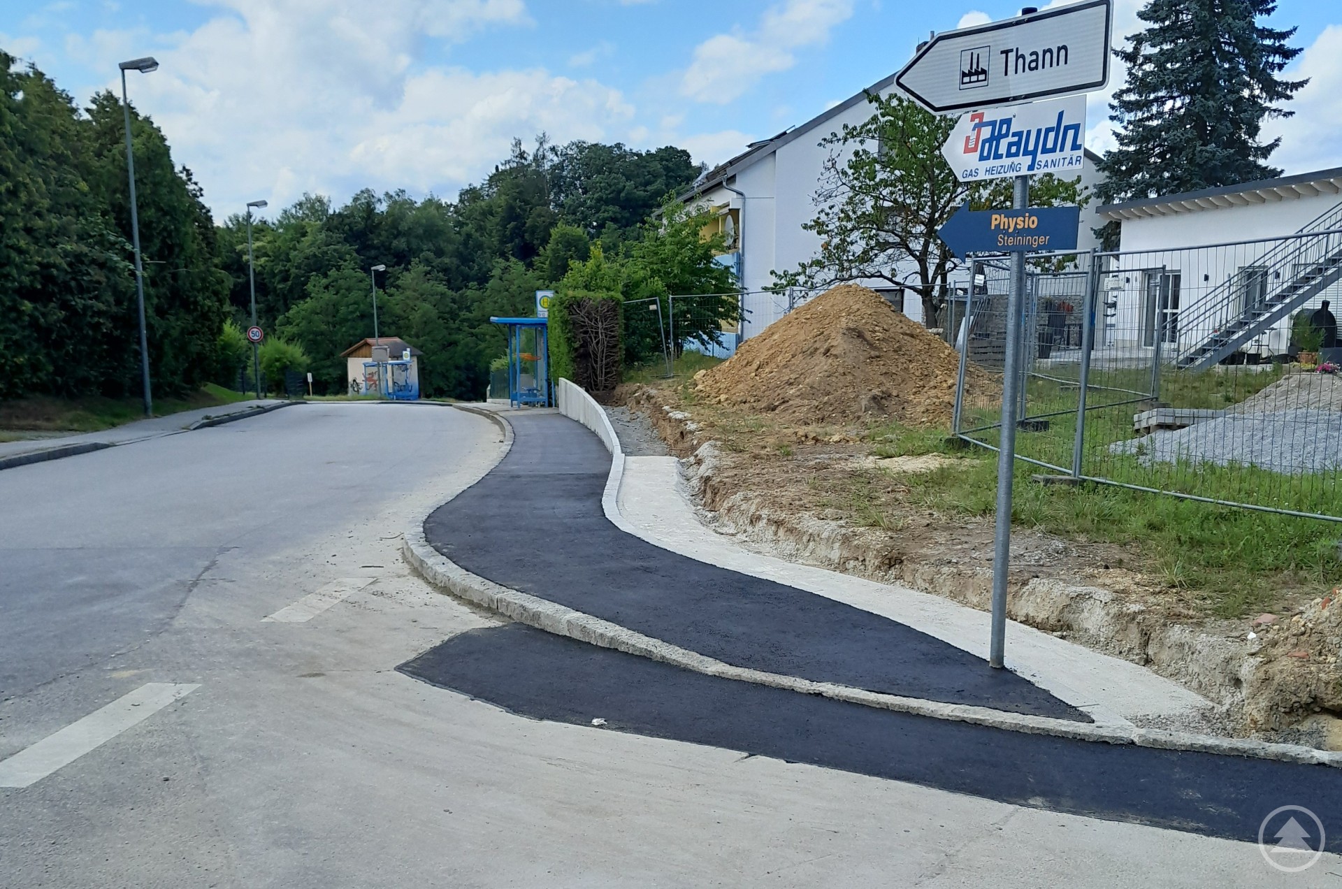 Neu errichteter Gehweg in der Hennebergerstraße Frisch asphaltierter Gehweg entlang einer Straße mit Straßenbeschilderung, im Hintergrund eine Bushaltestelle und ein Gebäude mit Baustelle.