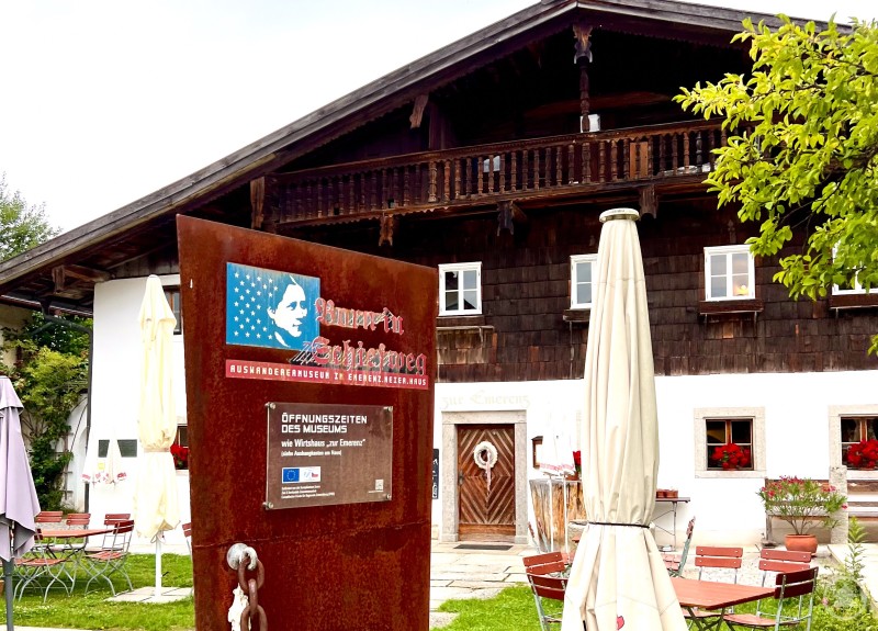 Das Auswanderermuseum „Born in Schiefweg“ mit dem Wirtshaus „zur Emerenz“. Außenansicht eines traditionellen bayerischen Bauernhauses mit Gastgarten, davor ein Schild mit der Aufschrift „Born in Schiefweg“.