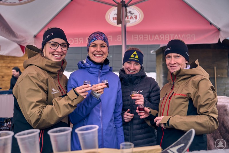 Gute Laune und geselliges Miteinander an den Ständen der Après-Ski-Party in Bodenmais. Vier Frauen stehen lachend an einem Getränkestand und stoßen mit kleinen Gläsern an. Sie tragen Winterjacken und Mützen, im Hintergrund ist ein Ausschankzelt zu sehen.