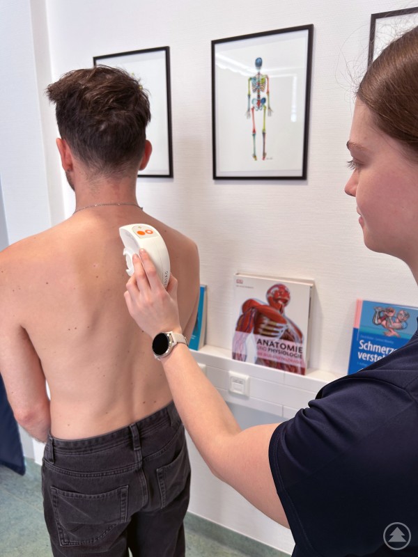 Mit einem handgeführten Messgerät wird die Wirbelsäule im Bereich des Rückens analysiert. Eine Mitarbeiterin führt bei einem Mann einen Rückenscan mit einem handgeführten Messgerät an der Wirbelsäule durch.