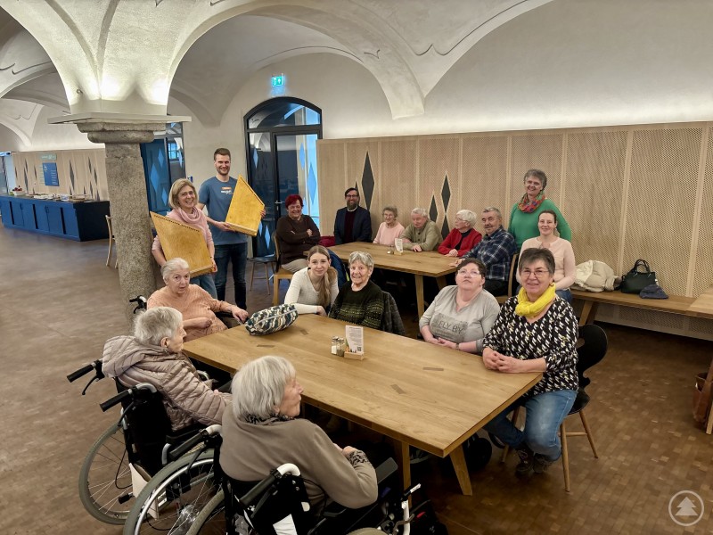 Voller Freude und mit voller Konzentration erzeugten die Teilnehmerinnen und Teilnehmer die ersten Klänge auf der Zauberharfe angeleitet und beobachtet von (hinten von links) Miriam Scholz und Dominik Hilgart (Volksmusikakademie), Andrea Obermeier (Rosenium Freyung, 2.v.r.) sowie Christian Fiebig (Landratsamt Freyung-Grafenau). Eine Gruppe von Seniorinnen und Senioren sitzt gemeinsam mit Betreuenden an mehreren Tischen in einem hellen Saal der Volksmusikakademie. Einige Teilnehmer halten Instrumente der sogenannten Zauberharfe in den Händen, während sie gemeinsam musizieren und sich austauschen.