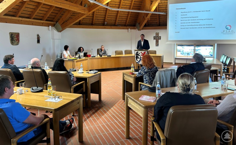 Landrat Sebastian Gruber begrüßte als Gastgeber die Mitglieder der Psychosozialen Arbeitsgemeinschaft (PSAG) Passau/Freyung-Grafenau im Landratsamt. Ein Mann spricht am Rednerpult im großen Sitzungssaal des Landratsamts Freyung. Um ihn herum sitzen zahlreiche Teilnehmende an Tischen, während auf einer Leinwand die Tagesordnung der PSAG-Mitgliederversammlung angezeigt wird.