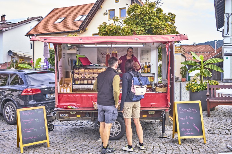 Am Stand des Käseparadieses Seiderer herrschte beim Auftakt des Bodenmaiser Wochenmarktes reger Andrang. Verkaufsstand mit rotem Wagen, beschrifteten Tafeln und Käseauswahl; zwei Kunden im Gespräch mit der Verkäuferin.