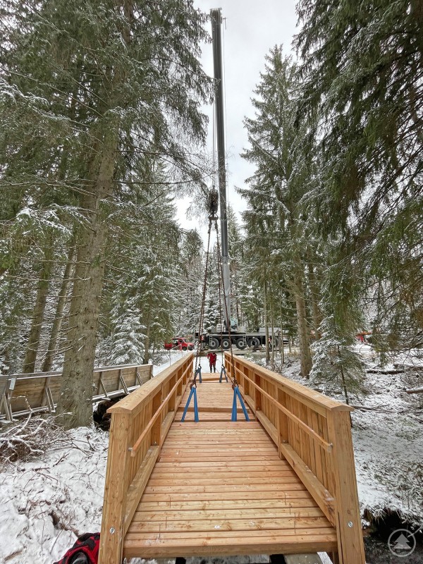 Noch am Haken: Die Brücke über die Schwarzach wurde in einem Stück eingehoben. Eine komplett vorgefertigte Holzbrücke wird mittels Kran durch einen verschneiten Wald gehoben. Die Brücke hängt an Stahlseilen, während Mitarbeitende der Nationalparkverwaltung sie auf die vorbereiteten Fundamente einweisen. Umgeben ist die Szene von schneebedeckten Bäumen und winterlicher Landschaft.