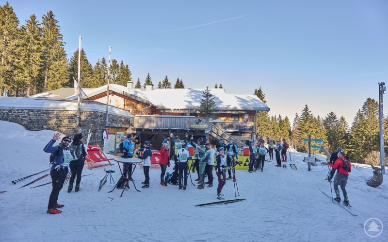 Zahlreiche Skilangl&auml;ufer machen Rast oder feiern gesellig vor der verschneiten Chamer H&uuml;tte. Im Vordergrund entspannte Sportlerinnen und Sportler mit Skiern, im Hintergrund die urige H&uuml;tte im Wald.