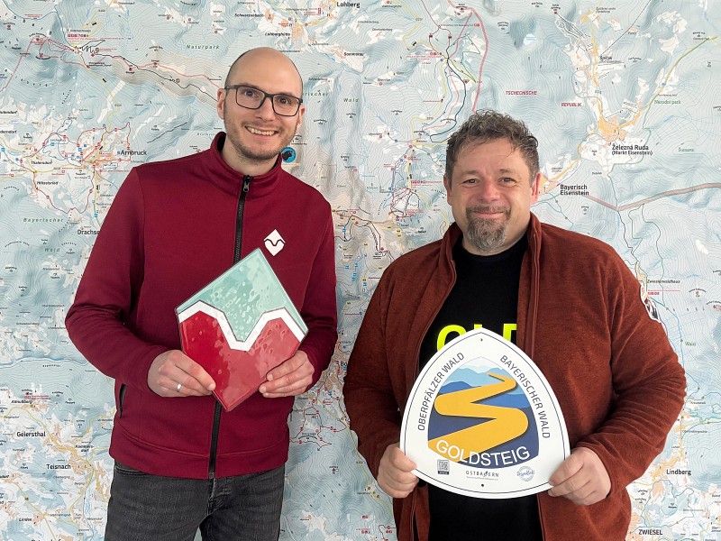 Bodenmais Tourismus und das Goldsteig-Team werben gemeinsam für die Publikumswahl zu Deutschlands schönstem Wanderweg 2026. Zwei Männer stehen vor einer großen Karte und halten Schilder zum Wildes-Wasser-Weg und zum Goldsteig in die Kamera.