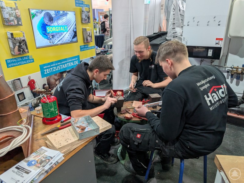 In Zusammenarbeit entsteht eine Rose aus Kupfer Drei Auszubildende sitzen im Kreis und formen mit verschiedenen Zangen eine Rose aus Kupfer.